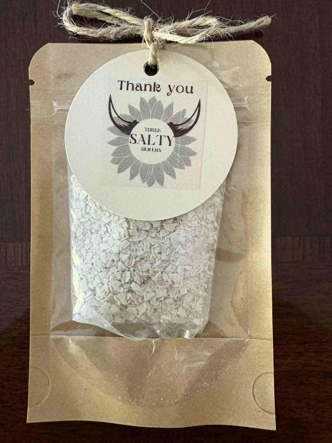 Kraft paper pouch filled with dehydrated sourdough starter flakes, sealed with a clear window and tied with rustic twine featuring a round “Thank You” tag labeled “Three Salty Heifers.”