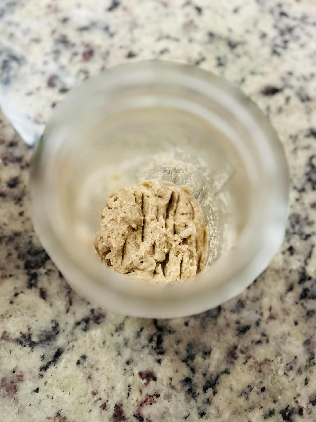 Close-up of rehydrated sourdough starter in a glass jar on a speckled countertop, showing a thick, bubbly texture as it begins to activate.