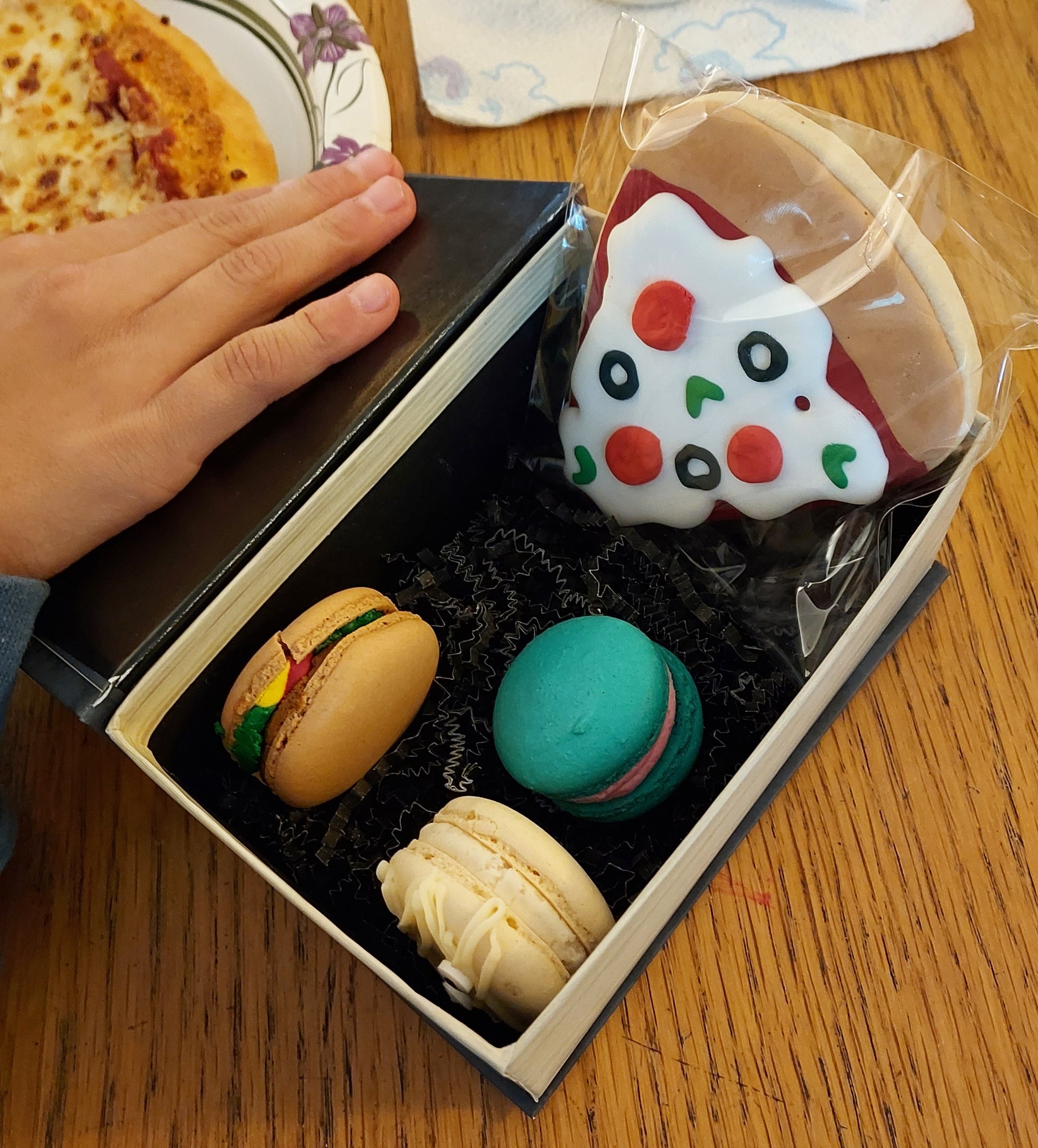 Box of colorful macaron cookies with a hand holding it on a wooden table.