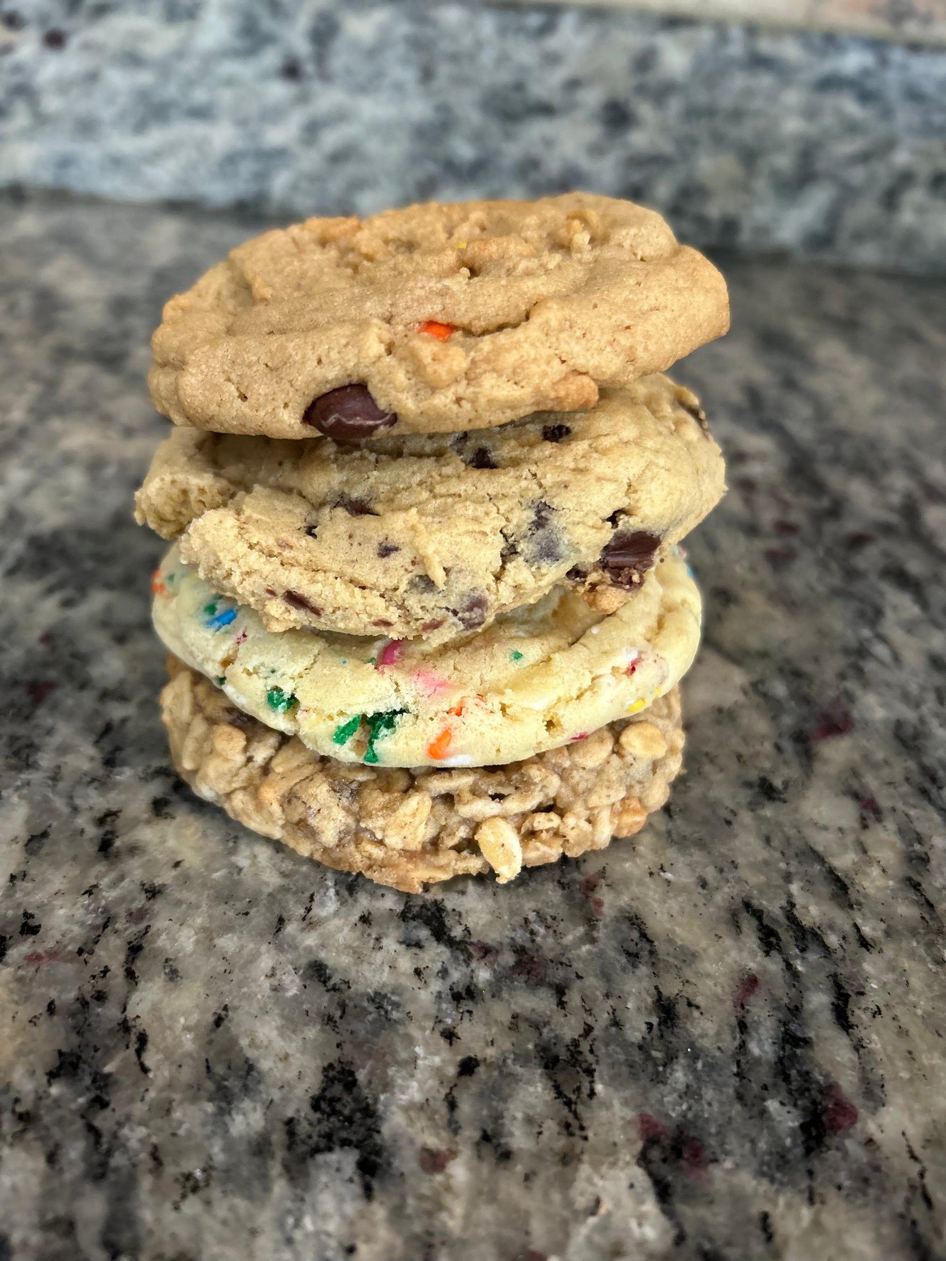Assorted gourmet cookies including chocolate chip, oatmeal raisin, sugar cookie, and birthday cake flavors, displayed in a box.