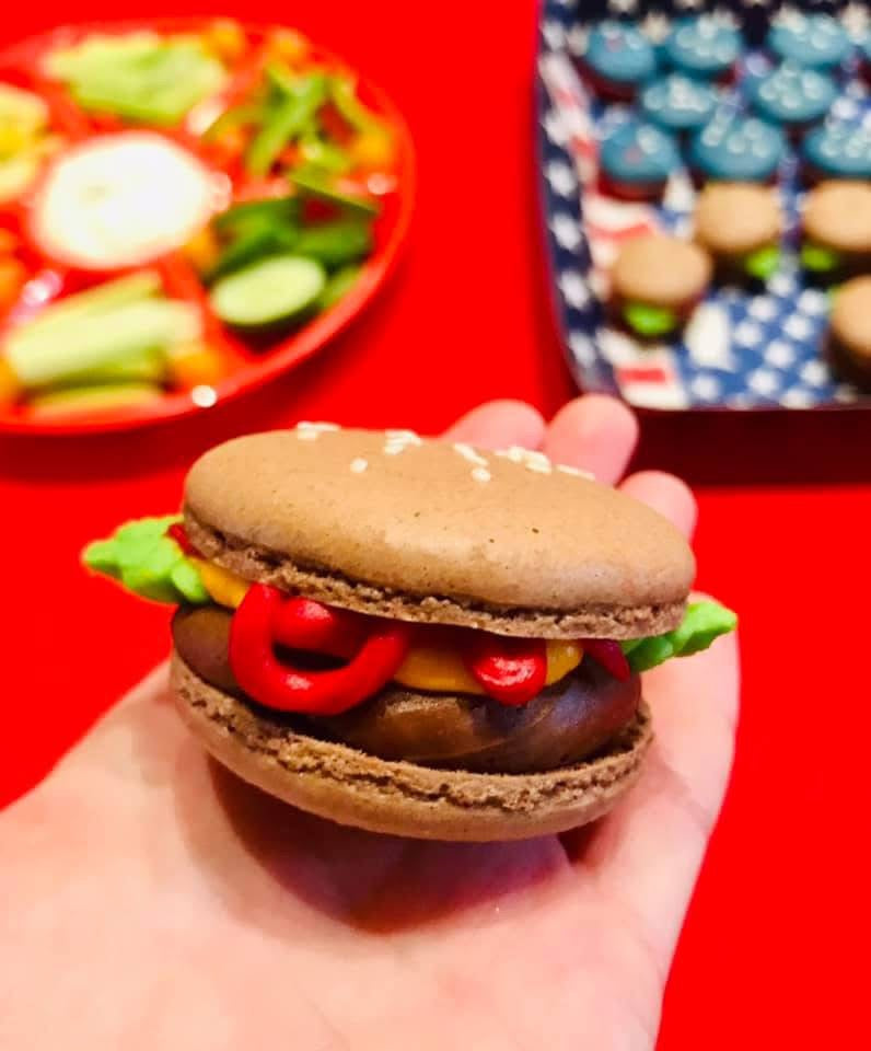 Close-up of a hand holding a creative macaron styled like a hamburger, with chocolate filling as the patty and colored icing resembling lettuce, ketchup, and mustard.