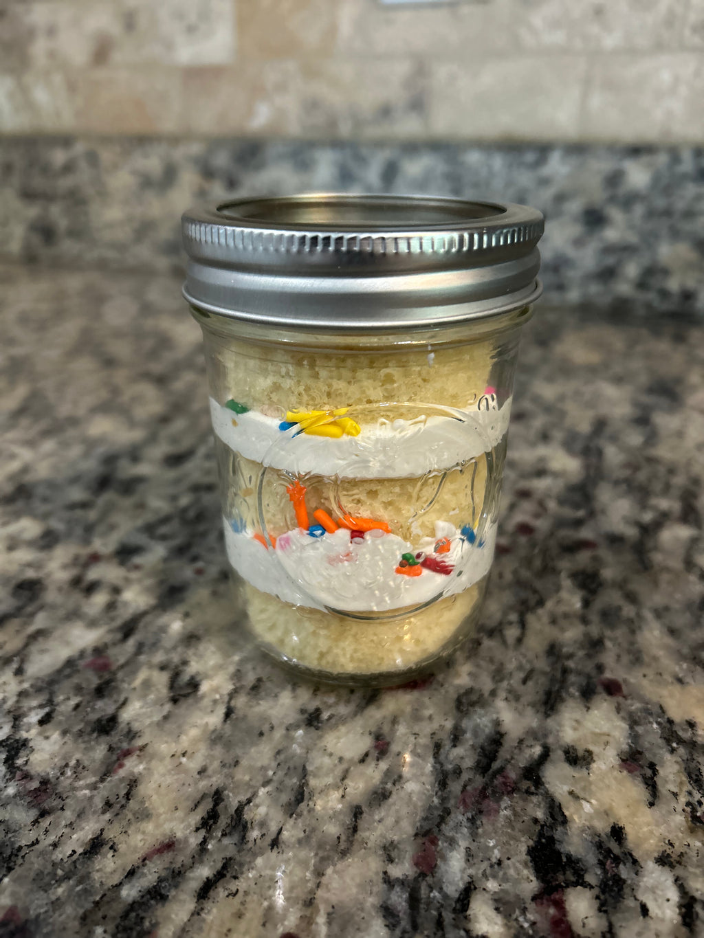 Layered cake in a mason jar with frosting and sprinkles, sealed with a lid and ribbon