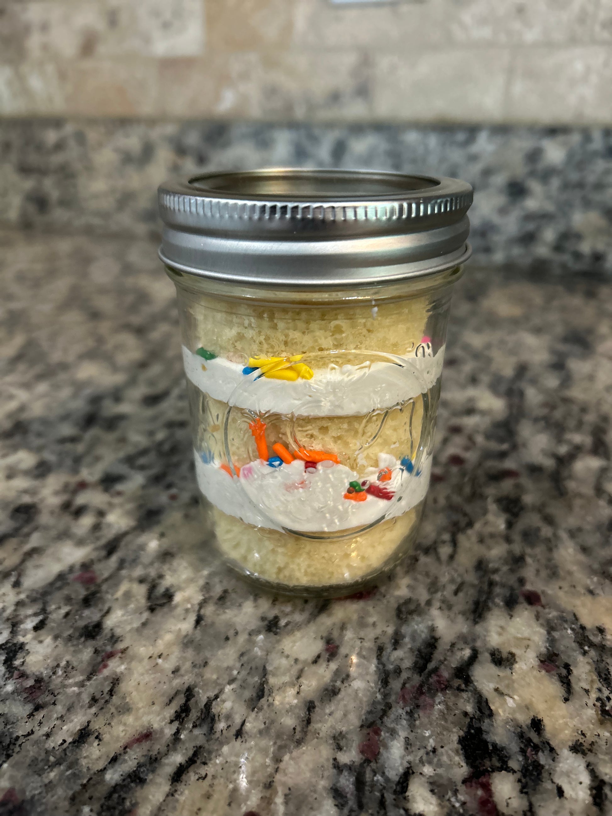 Layered cake in a mason jar with frosting and sprinkles, sealed with a lid and ribbon