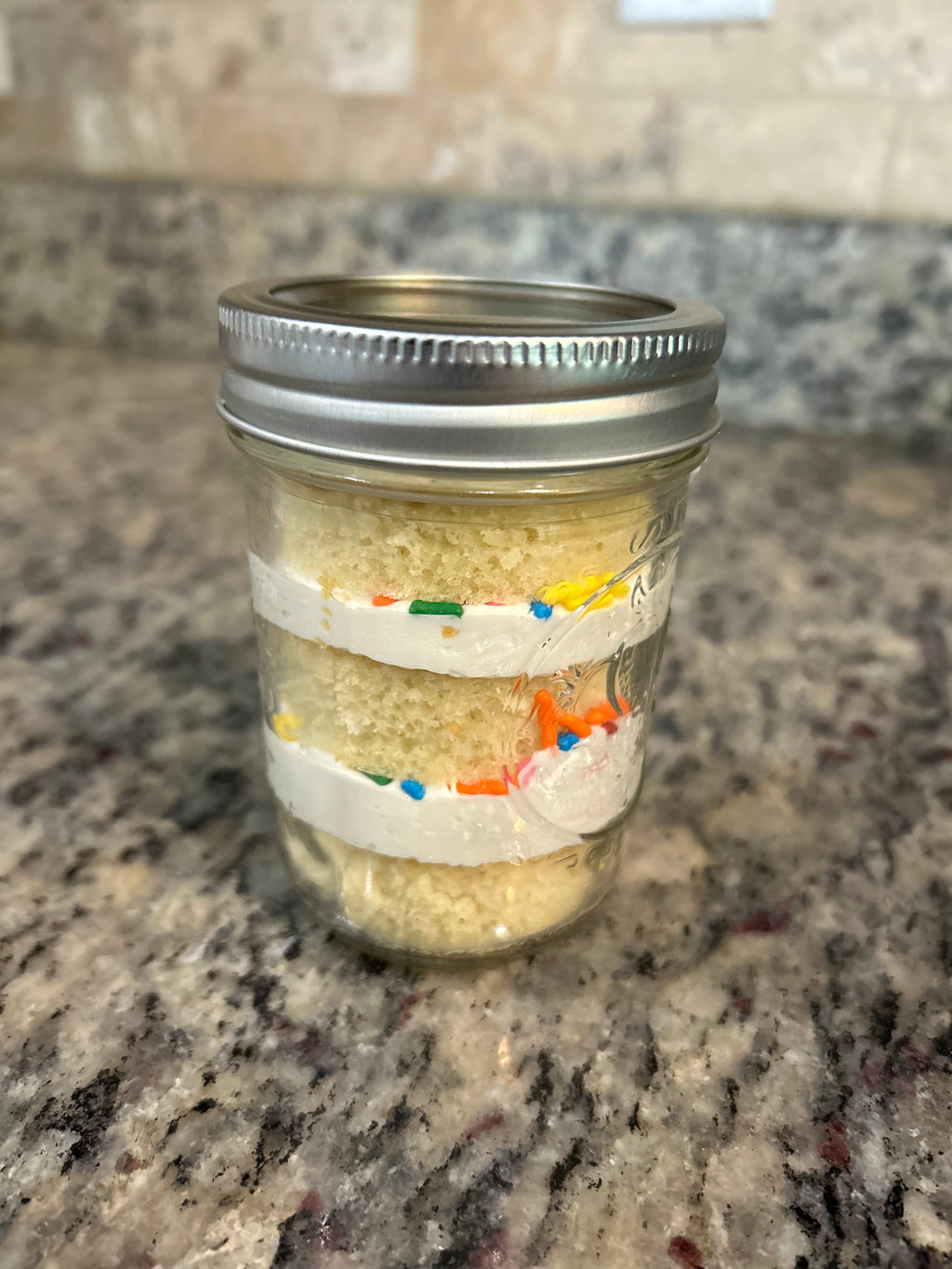 Layered cake in a mason jar with frosting and sprinkles, sealed with a lid and ribbon