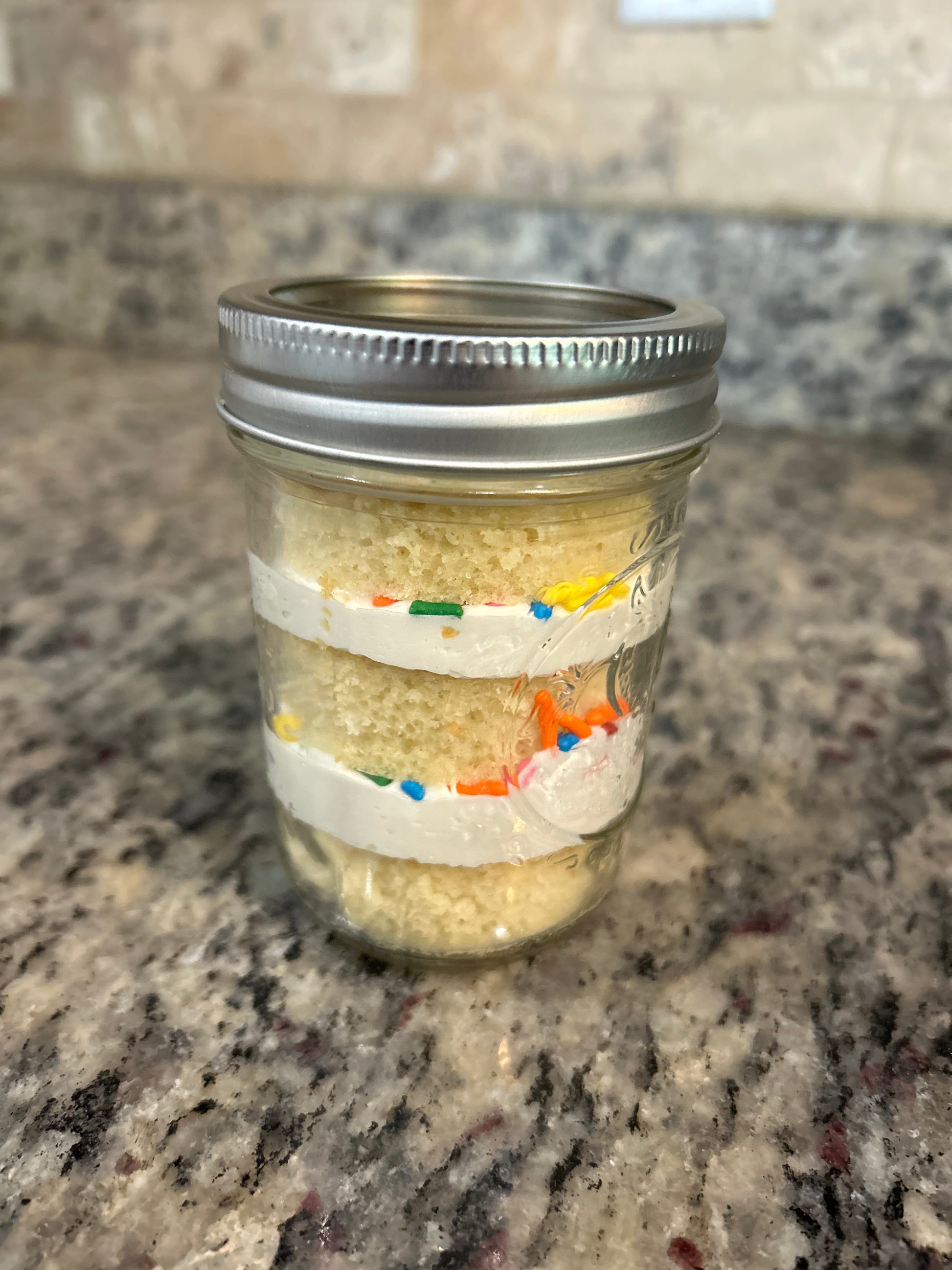 Layered cake in a mason jar with frosting and sprinkles, sealed with a lid and ribbon