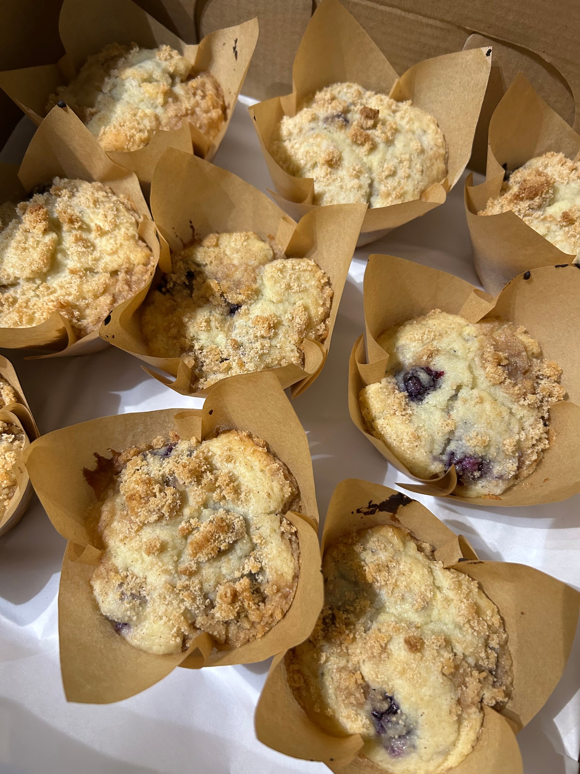 Freshly baked blueberry muffins with crumb topping, wrapped in brown tulip-style parchment liners, arranged in a bakery box.