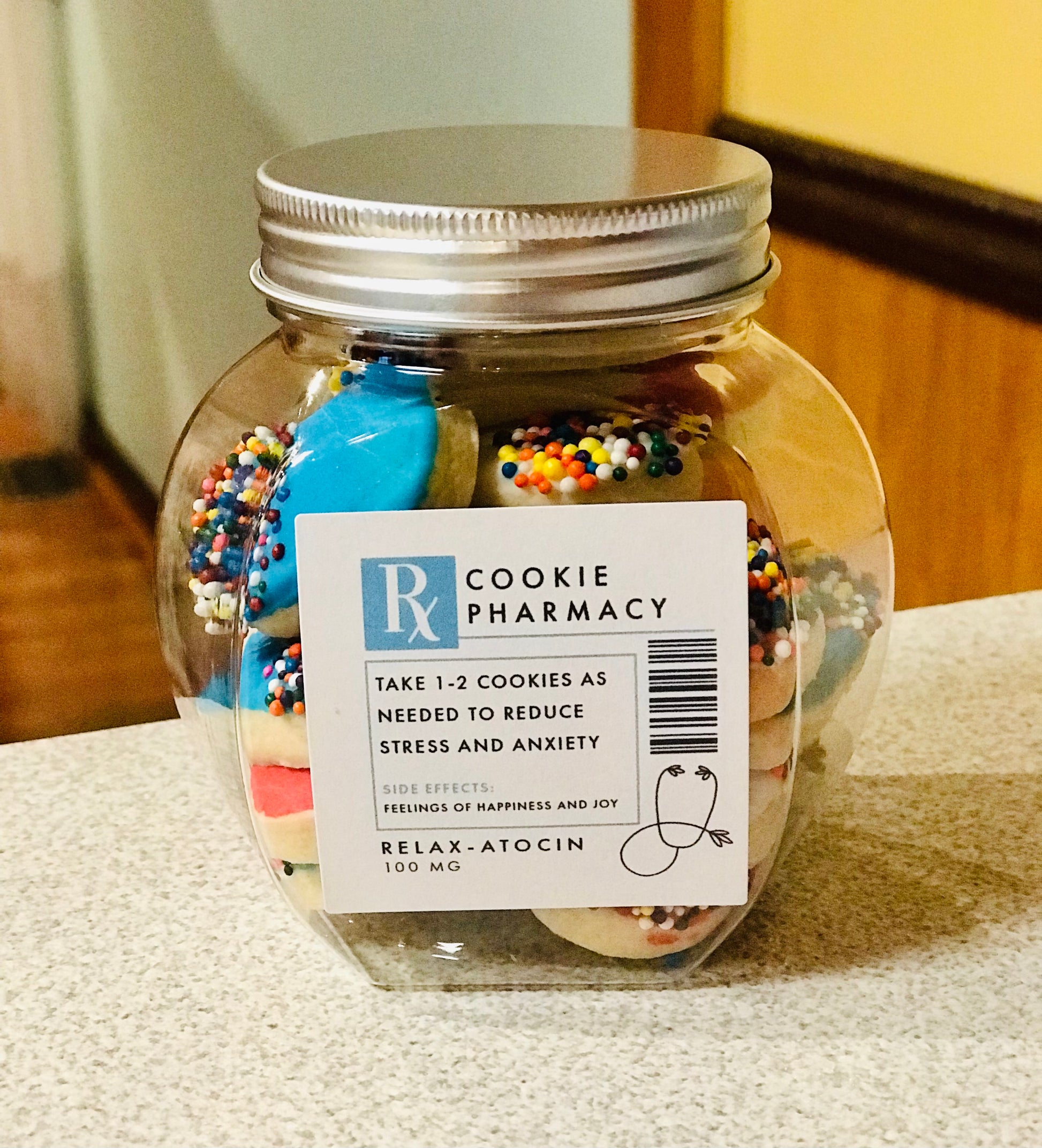 Clear glass jar labeled ‘Cookie Pharmacy’ filled with colorful frosted cookies topped with rainbow sprinkles. The label humorously mimics a prescription, suggesting to take 1–2 cookies as needed to reduce stress and anxiety, with side effects including happiness and joy.
