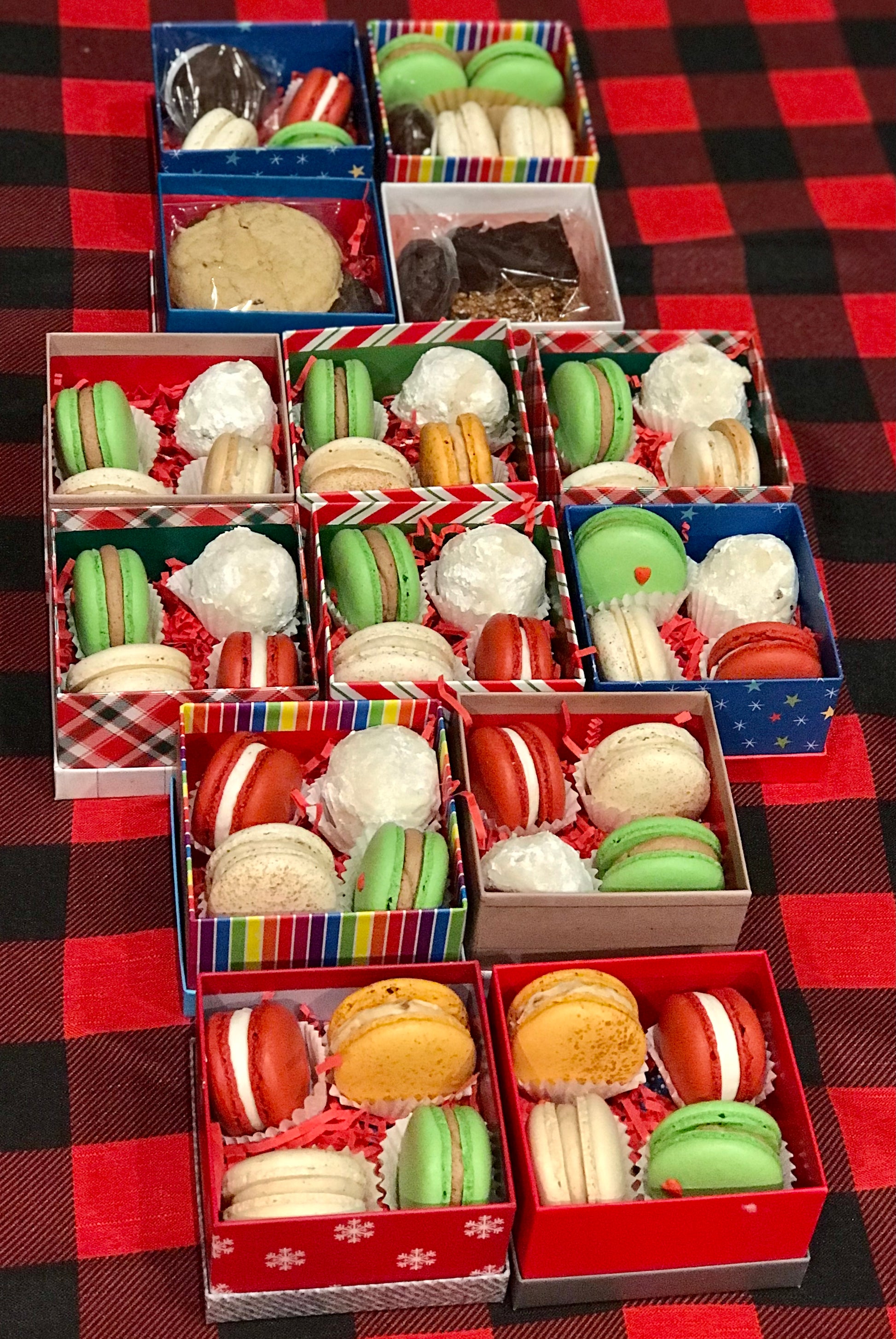 Assorted holiday cookie and macaron gift boxes displayed on a red and black plaid tablecloth. Each box includes colorful macarons in red, green, white, and yellow, paired with powdered snowball cookies, brownies, or classic cookies, arranged in festive packaging with red crinkle paper.