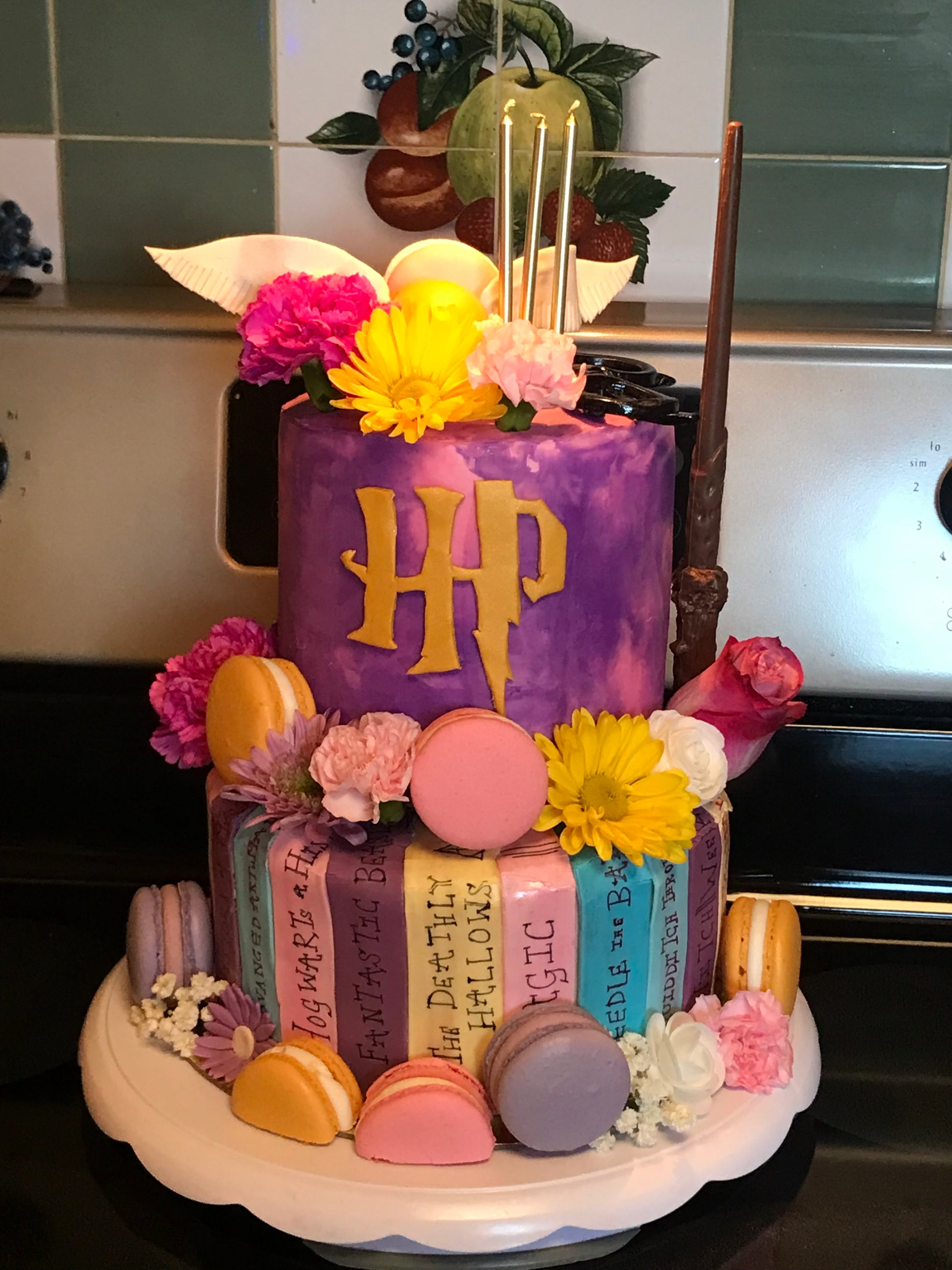 Two-tier Harry Potter-themed cake decorated with fresh flowers, macarons, golden candles, and fondant books referencing magical titles. The top tier features a gold HP logo, golden snitch wings, and a chocolate wand.